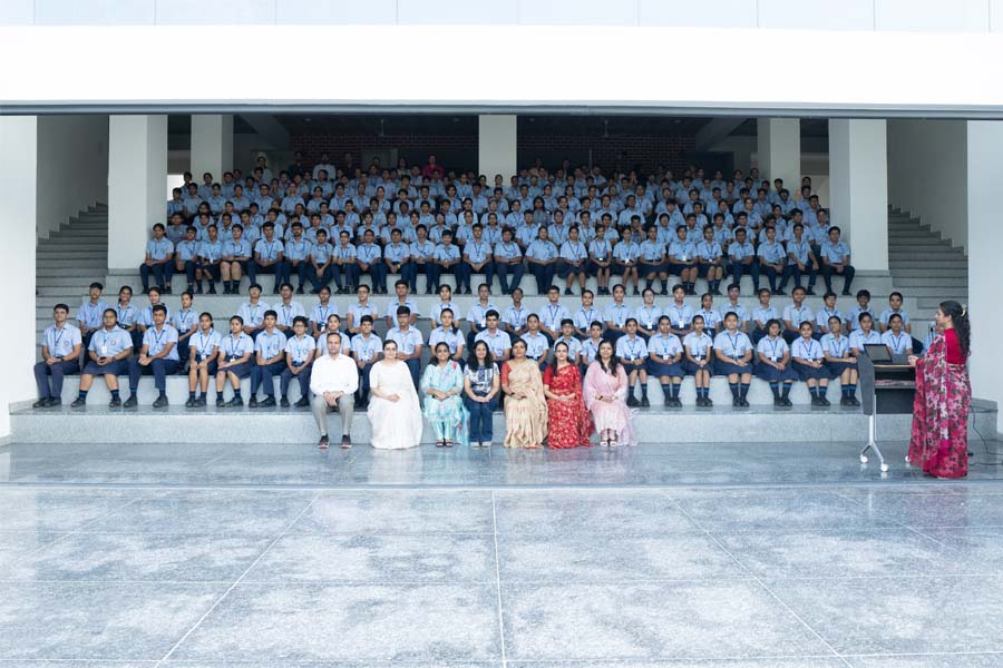 A large group of students in uniforms is seated in bleachers for a group photo in front of a building, with seven adults seated in front and two standing at a podium, all within the open spaces of the campus.