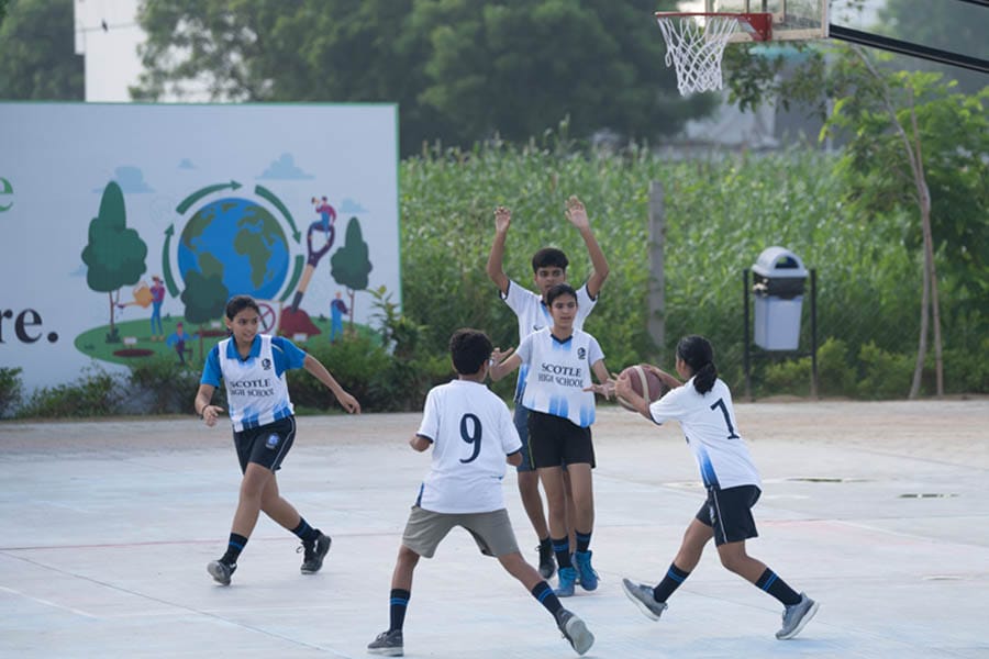 Five children are playing basketball on an outdoor court. Four wear white jerseys, and one wears a black jersey. A mural with a globe and children in the background highlights the spirit of sports. Trees and bushes are visible, adding a touch of nature to the vibrant scene.