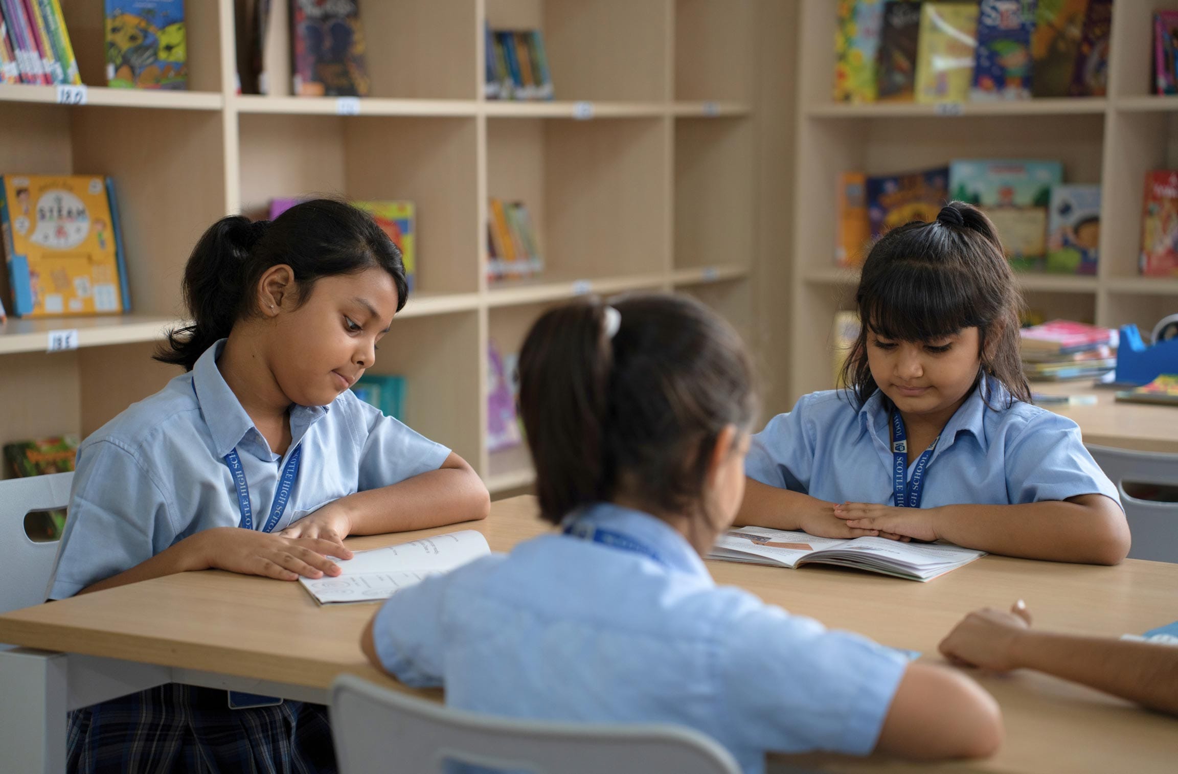 Three young students in school uniforms sit at a table reading books in a classroom with bookshelves in the background.