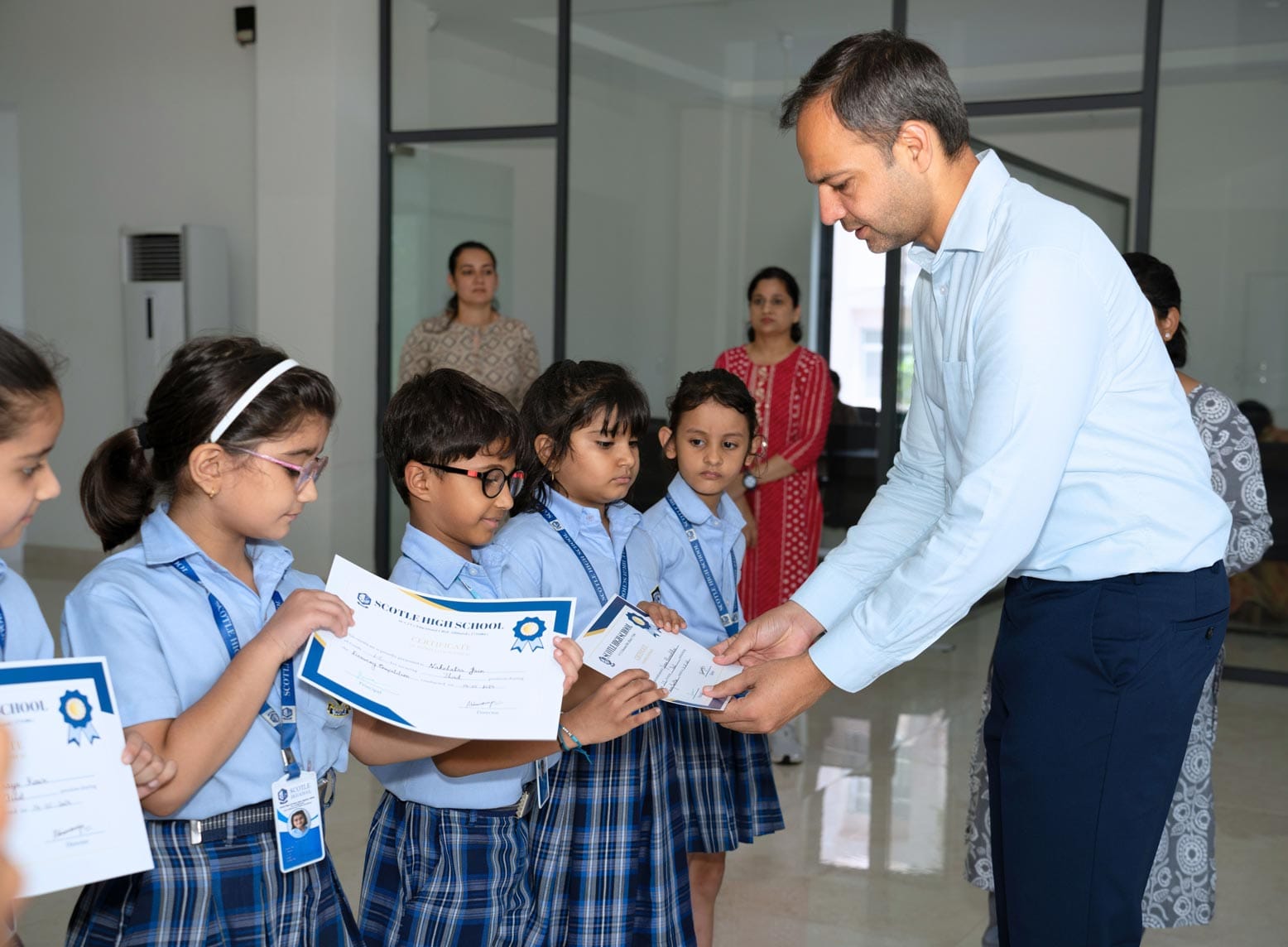 A man in a blue shirt hands out certificates to a group of young students in school uniforms. Other children and adults watch in the background.