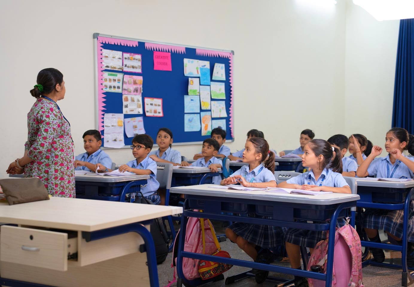 A teacher stands in front of a classroom of attentive students in uniform, with drawings and posters pinned on a bulletin board behind her. The students are seated at desks in rows.