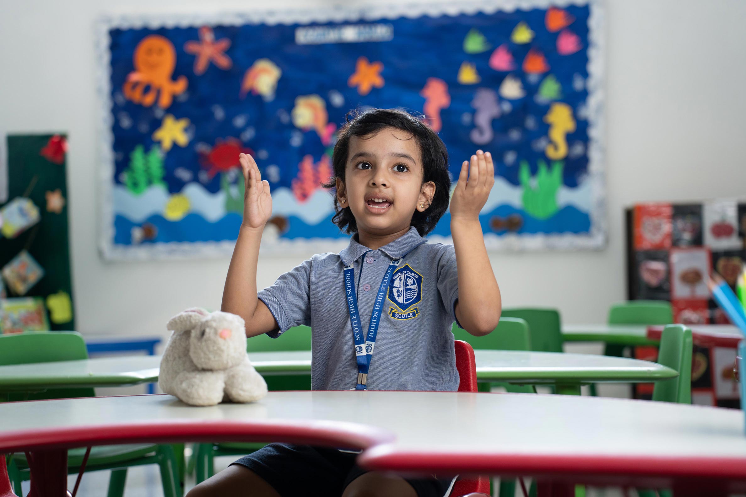 A young child sits at a classroom table with a plush toy in front, gesturing with hands. The background features a colorful underwater-themed bulletin board.