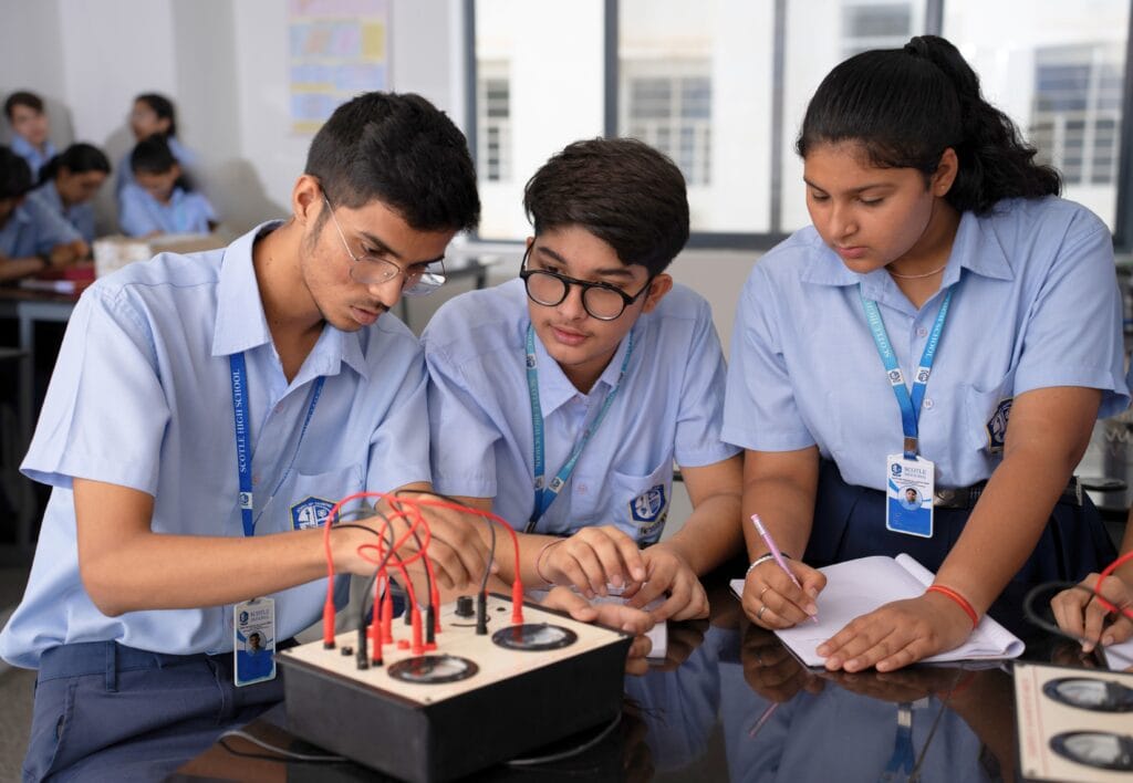 Three high school students in uniforms conduct an experiment with an electrical circuit board on a desk in a classroom. One student takes notes while the other two adjust the wires on the circuit.