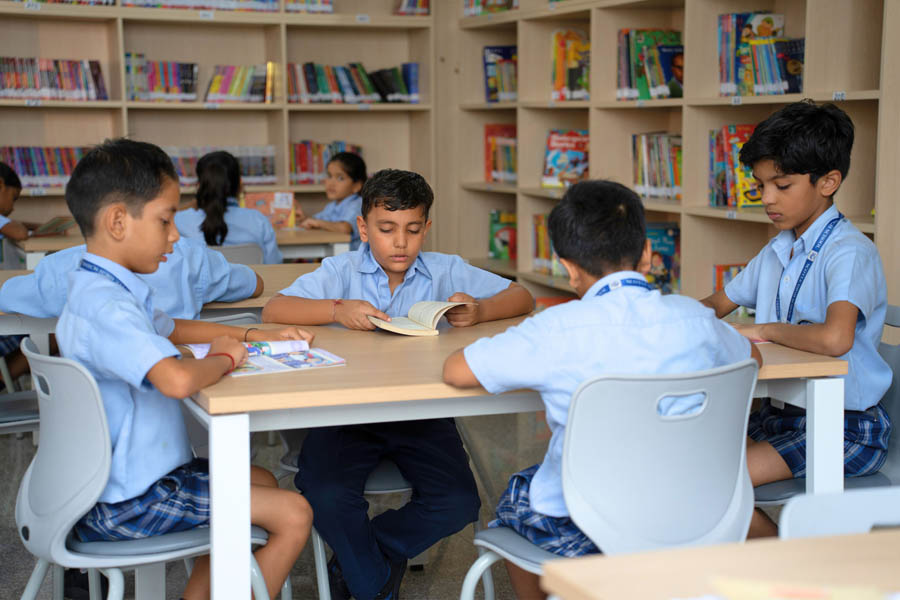 Children in school uniforms are sitting at tables and reading books in a library, surrounded by the well-organized infrastructure of shelves lined with colorful books.