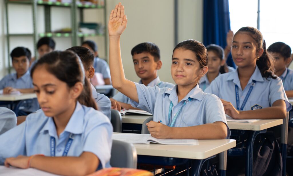 A middle school classroom with students in uniforms seated at desks, one student raising their hand while others attend to their work.