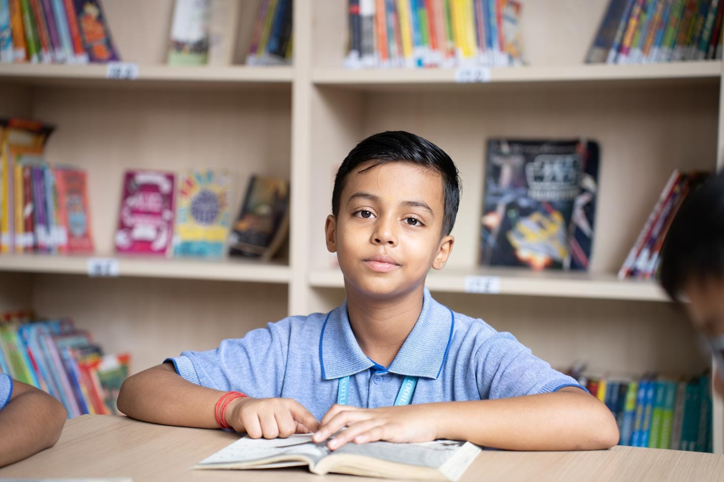 A young boy in a blue shirt is sitting at a table with an open book in a library. Shelves filled with colorful books are visible in the background.