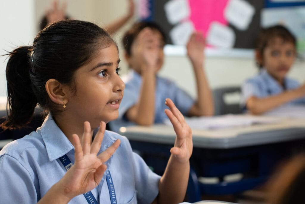 A young girl in a primary school uniform raises her hand in a classroom, with other children in similar attire raising their hands in the background.