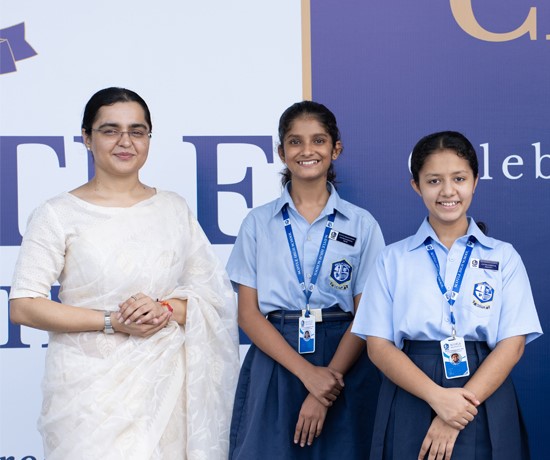 A woman in a white saree stands next to two smiling girls in blue school uniforms, posing in front of a banner displaying the Principal's Message.