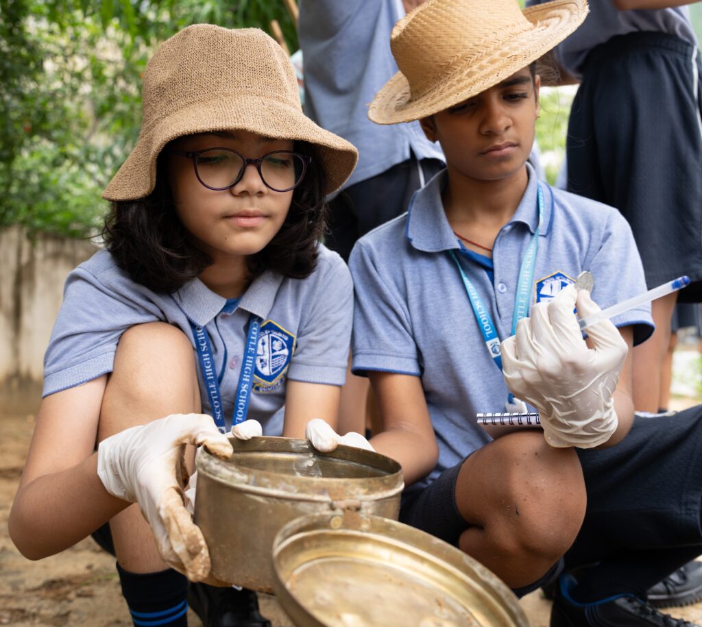 Two students wearing hats and gloves examine a container and make notes outdoors, showcasing their school uniforms with emblems on their shirts. The activity fosters a strong sense of community and learning among the group.