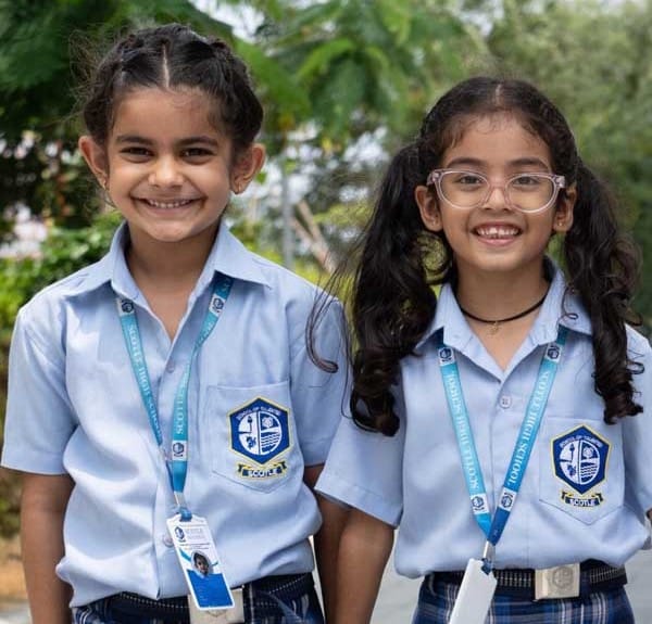 Two young girls in school uniforms, wearing ID cards and lanyards, stand side by side outdoors, smiling at the camera.